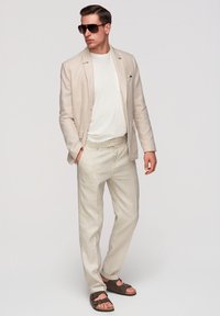 Homme portant un costume beige, un t-shirt blanc, des lunettes de soleil foncées et des sandales marron, debout avec une main dans la poche devant un fond uni.