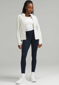 Jachetă sport albă cu fermoar, design cu gât înalt, asortată cu colanți bleumarin și adidași albi. Prezintă țesături elegante, netede și o formă ajustată.