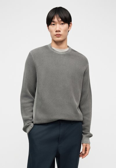 Νέος άνδρας με μαύρα μαλλιά, φορώντας υφασμάτινο γκρι πουλόβερ με στρογγυλή λαιμόκοψη και σκούρο μπλε παντελόνι, στέκεται έχοντας το ένα χέρι στην τσέπη.