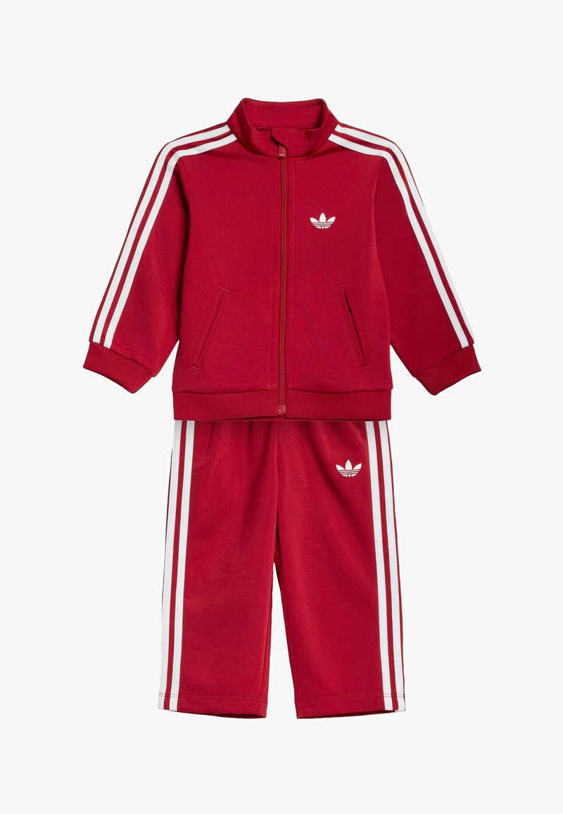 Červená dětská tepláková souprava s bílými pruhy na rukávech a kalhotách, bunda na zip a loga Adidas trefoil na hrudi a kalhotách.