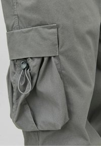 Pantalon cargo gris avec une poche latérale comportant un détail de tissu froncé et une fermeture par cordon de serrage, fabriqué dans un matériau doux et texturé.