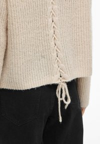 Pull tricoté beige clair avec un détail tressé le long du dos et un cordon de serrage à l'ourlet, associé à un jean noir.