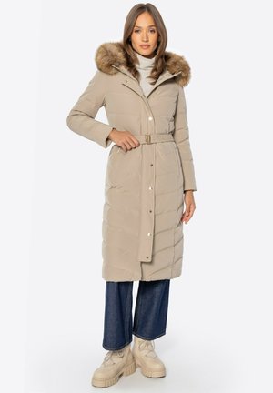 Femme portant un long manteau matelassé beige avec une ceinture à la taille et une capuche en fausse fourrure, un jean large bleu et des bottes beiges épaisses, debout.