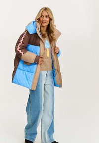 Žena v pestrofarebnej modro-beige-hnedej prešívanej bunde cez béžový sveter so vzorom huby, v širokých dvojfarebných džínsach a čiernych čižmách.