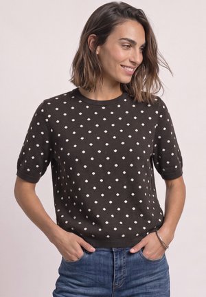 Femme souriante portant un haut noir à manches courtes avec des pois blancs et un jean bleu, mains dans les poches sur un fond clair uni.