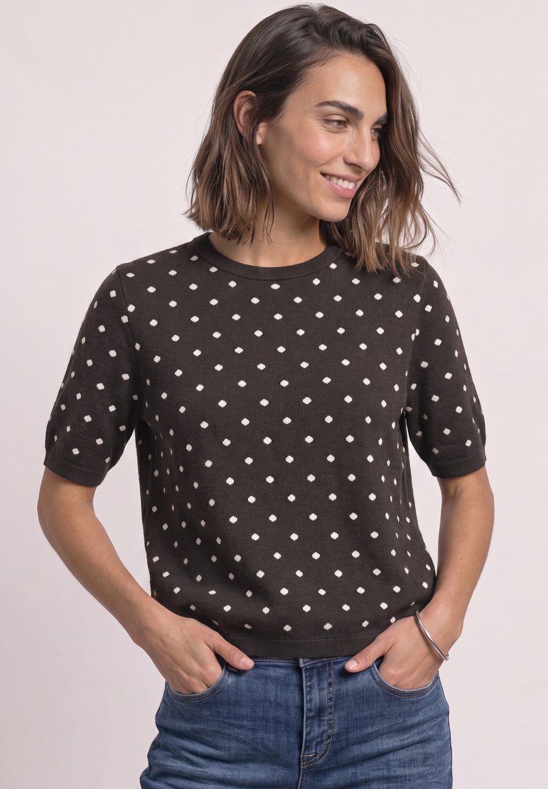 Femme souriante portant un haut noir à manches courtes avec des pois blancs et un jean bleu, mains dans les poches sur un fond clair uni.