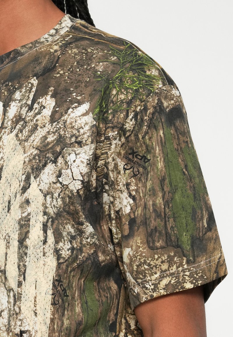 Gros plan sur une personne portant une chemise à manches courtes avec un motif camouflage aux tons terreux et des détails en feuillage vert, épaule et bras supérieur visibles.