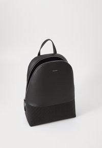 Fekete bőr hátizsák lekerekített tetejével, sima textúrával, ezüst cipzárral és mintás fekete alappal. Calvin Klein márkajelzése az elején.