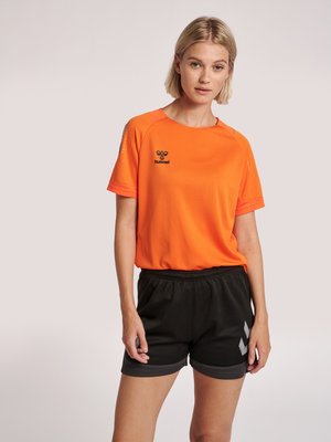 Mujer con camiseta deportiva naranja con logo negro y pantalones cortos deportivos negros con patrón de cheurón gris sobre fondo claro.