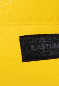 Material impermeable de color amarillo con gotas, presenta una etiqueta rectangular negra que muestra "EASTPAK" con un diseño de globo.