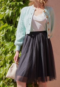 Ljust blått stickat cardigan med kabelmönster och guldfärgad knappar, kombinerat med en figurnära svart tylskjol. Modellen håller en liten vit handväska.