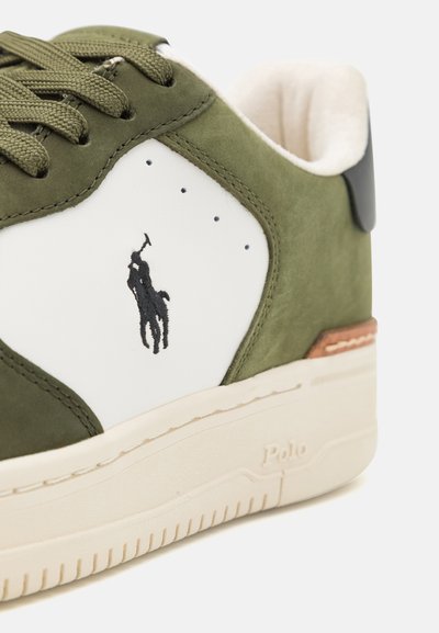 Zaļi zamšādas sporta apavi ar baltu ādas sānu paneli, uz kura ir melns polo logos. Akcentēti ar beigu gumijas zoli un brūnu šuvju detaļām.