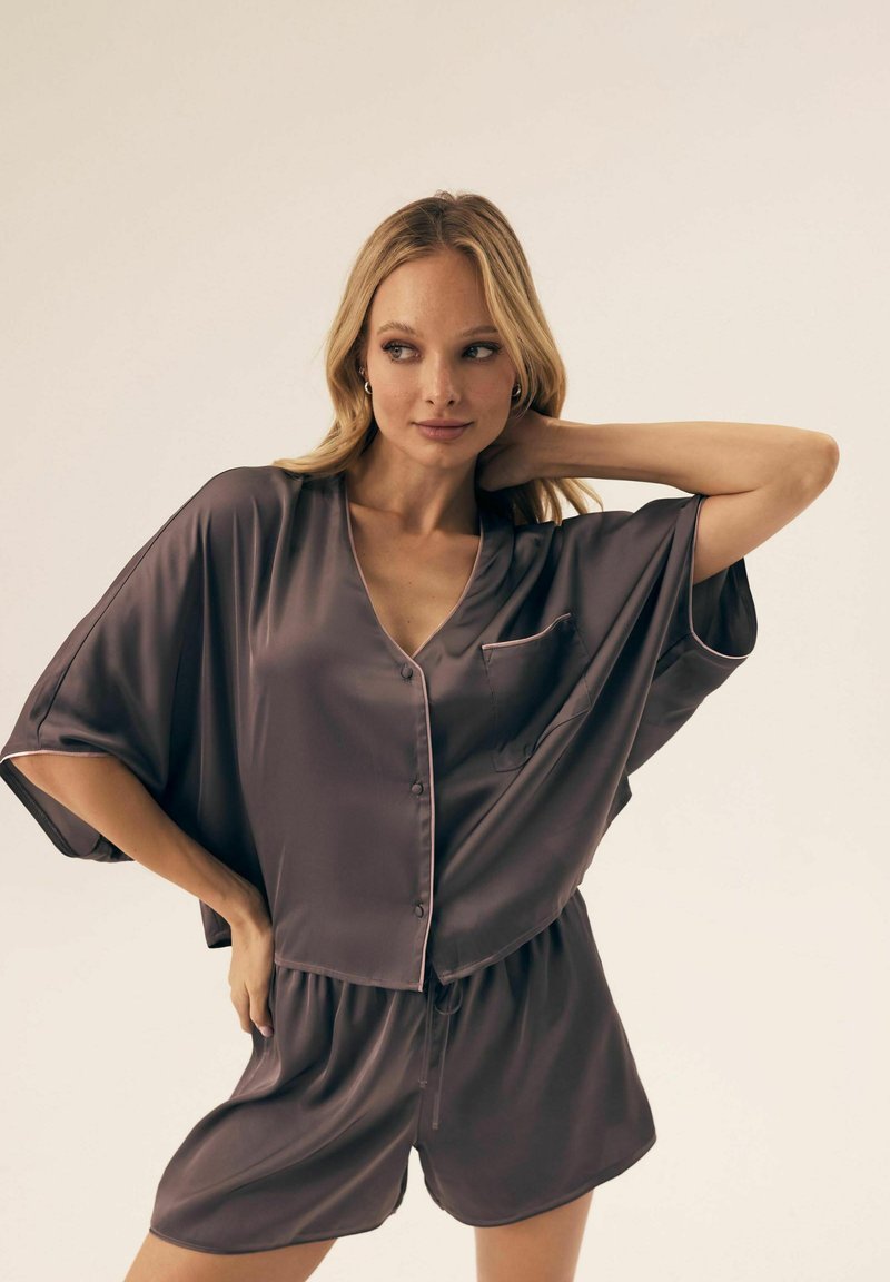 Blondynka pozująca z ręką za szyją, ubrana w luźną, jedwabistą, ciemnoróżową koszulę na guziki i dopasowane szorty na jednolitym tle.