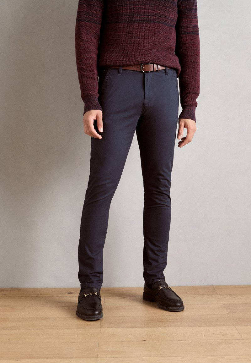 Granatowe spodnie slim-fit wykonane z bawełny, w połączeniu z burgundowym swetrem z dzianiny. Brązowy skórzany pasek i czarne buty z złotymi akcentami.