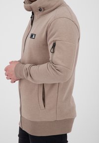 Uomo in felpa a zip beige con colletto alto, polsini a costine, tasche laterali e una tasca con zip sulla parte superiore del braccio, in piedi di lato.