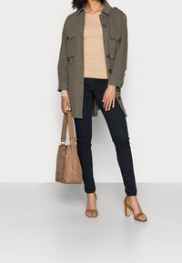 Nainen, jolla on yllään oliivinvihreä nappijakku, beige ribbed-neule, mustat housut, vaaleanruskeat korkokenkäsaappaat, ja hän pitää vaaleanruskeaa käsilaukkua sivussa.