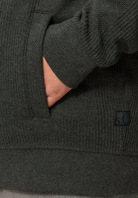 Dunkelgrüner Strickpullover mit gerippter Textur, einer Vordertasche und einem kleinen Markenlabel. Die Bündchen zeigen ein ähnliches geripptes Design.