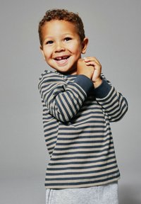 Uśmiechnięte małe dziecko z kręconymi włosami, mające na sobie paskowaną koszulę z długim rękawem, trzymające dłonie blisko twarzy na jednolitym tle.