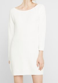 Donna che indossa un abito lungo in maglia bianca a maniche lunghe con polsini e orlo a costine, in piedi davanti a uno sfondo chiaro e uniforme.