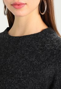 Schwarzer, strukturierten Pullover mit Rundhalsausschnitt. Nahaufnahme zeigt dicken, flauschigen Stoff und kleine graue Fasern. Große silberne Creole sichtbar.