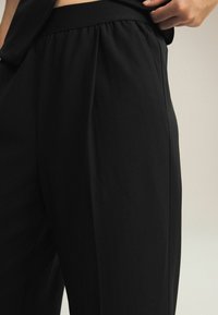 Schwarze weite Hosen mit elastischem Bund, die eine glatte Textur und eine elegante Silhouette aufweisen, gezeigt auf Taillen- und Oberschenkelhöhe.
