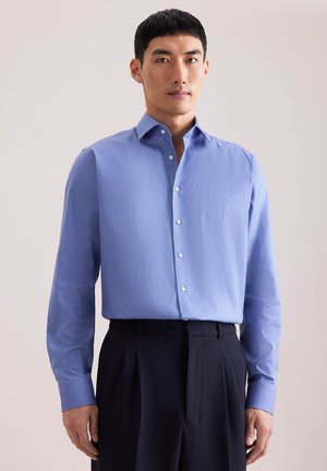 Man draagt een lichtblauw patroon overhemd met knopen en lange mouwen, ingestopt in zwarte geplooide broek, tegen een effen achtergrond.