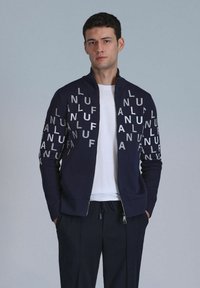 Navy zip-up sweatshirt met een wit logo op de voorkant. Heeft een staande kraag, lange mouwen en geribde manchetten. Gecombineerd met een wit T-shirt.