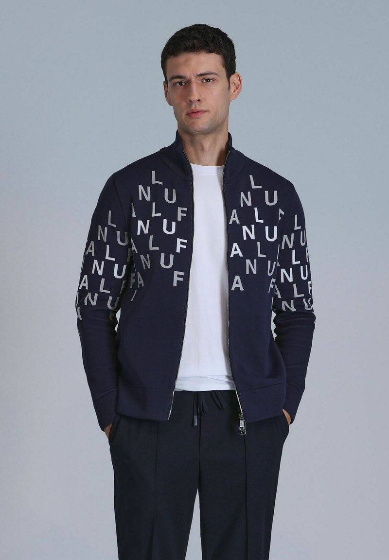 Navy zip-up sweatshirt met een wit logo op de voorkant. Heeft een staande kraag, lange mouwen en geribde manchetten. Gecombineerd met een wit T-shirt.