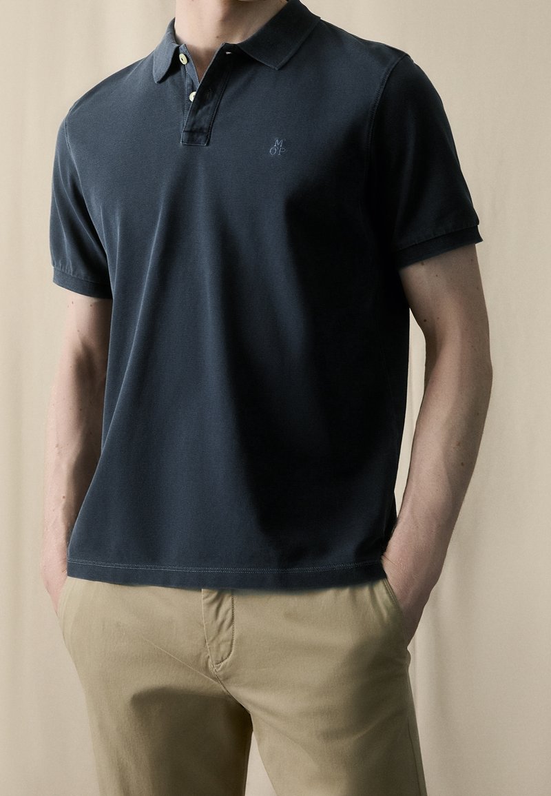 Homme portant un polo manches courtes bleu marine foncé avec boutons et pantalon beige, mains dans les poches, sur un fond neutre uni.