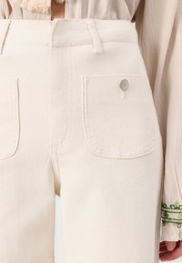 Pantalones beige de talle alto con bolsillos frontales y detalle de botón metálico, combinados con una blusa de manga larga color crema con bordado verde en el puño.