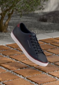 Baskets Tommy Hilfiger noires et rouges avec semelle blanche, lacets attachés, flottant au-dessus d'un sol en dalles de bois et de gravier, avec un arbre en arrière-plan.