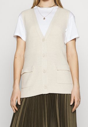 Gilet a coste beige con scollo a V, quattro bottoni e due tasche frontali, indossato sopra una maglietta bianca e una gonna verde oliva plissettata.