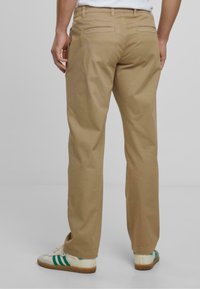 Pantaloni din bumbac bej, cu croială dreaptă, dotati cu două buzunare pe spate și o textură netedă. Purtați cu încălțăminte albă și dungi verzi.