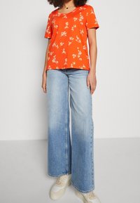 Orange kortærmet T-shirt med hvidt blomsterprint, parret med lyseblå vidde jeans og lyse sneakers.