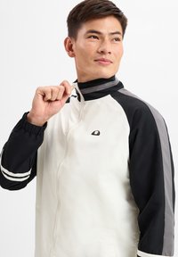 Chaqueta blanca y negra con detalles en gris, cierre frontal, cuello alto y puños elásticos. Presenta rayas en las mangas y un logotipo en el pecho.