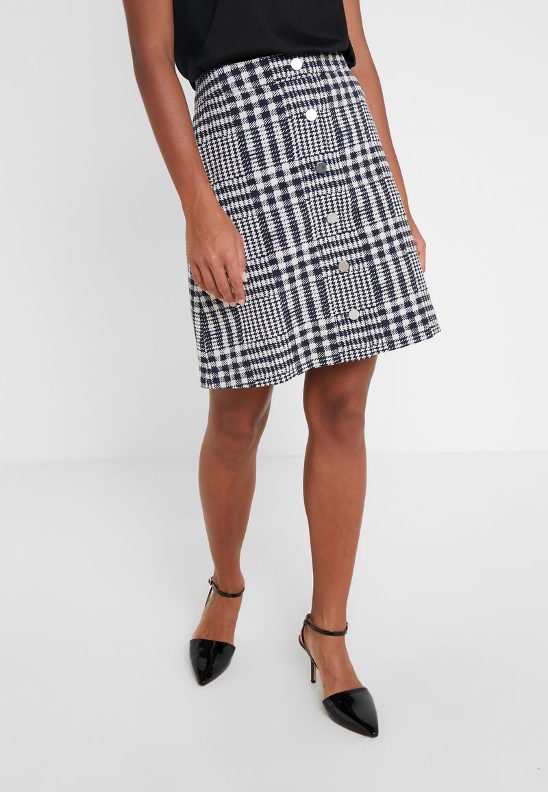 Femme portant une jupe à boutons devant en pied-de-poule noir et blanc avec des talons noirs à bout pointu et bride à la cheville, debout sur un sol blanc uni.