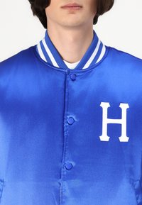 Jachetă din satin albastru cu un logo alb "H", închidere cu nasturi și guler ribbed cu dungi în alb și albastru. Textură netedă.