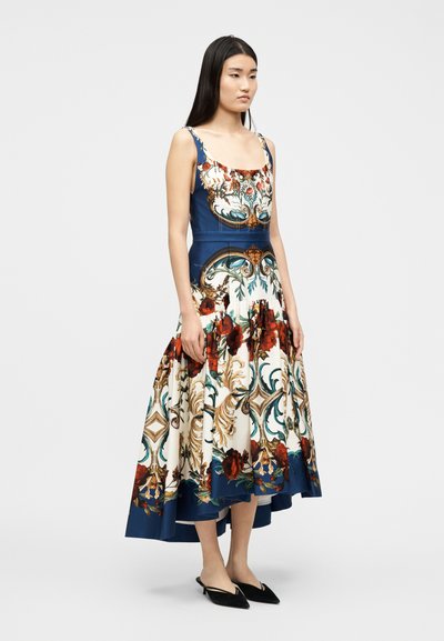 Femme portant une robe midi sans manches à motifs floraux et ornementaux, avec un ourlet asymétrique et des chaussures slingback noires à bout pointu.