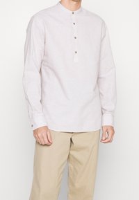 Ljusrosa långärmad skjorta med ståkrage, försedd med en frontknäppning med tre bruna knappar och ärmslut med rynkning.