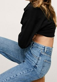 Personne portant un pull court noir et un jean taille haute bleu clair, assise de côté sur un tabouret en bois devant un fond uni.