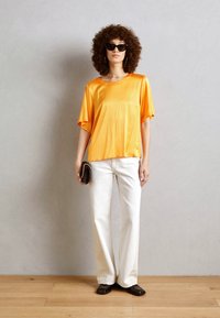 T-shirt arancione in satin con vestibilità ampia e maniche corte, abbinato a pantaloni bianchi a gamba dritta e occhiali da sole neri.