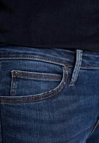 Blå denimjeans med en klassisk design, med kontrasterande orange sömmar och en framficka med silverdetaljer.