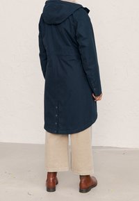 Granatowy długi płaszcz z kapturem, o gładkiej fakturze, z zapięciem na guziki przy mankietach oraz lekko zaokrąglonym dołem, noszony na beżowych szerokich spodniach.