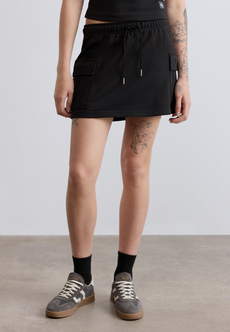 Μαύρη φούστα από τζέρσεϊ με ελαστική μέση και κορδόνι, δύο πλαϊνές τσέπες και άνετη εφαρμογή, συνδυασμένη με γκρι αθλητικά παπούτσια και μαύρες κάλτσες.