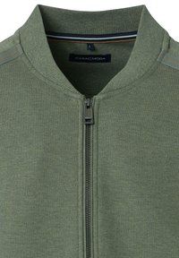 Olijfgroene zip-up jack met een geribbelde kraag, textuur stof en een voorkant rits. Heeft een subtiele gestreepte detail nabij de kraag.