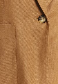 Bruine gestructureerde stof met een close-up van een op maat gemaakte jas met een zichtbare zak en een ronde, donkere knoop.