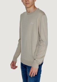 Maglione beige a maglia con maniche lunghe, scollatura rotonda e polsini a coste. Presenta un piccolo logo ricamato sul petto. Indossato con jeans blu.