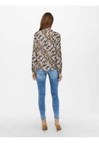 Blusa floreale con maniche lunghe e scollatura a chiave, abbinata a jeans skinny blu sbiaditi e tacchi nudi, su sfondo bianco.