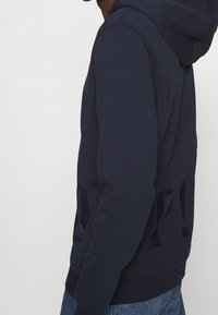 Man med en mörkblå hoodie med stora, delvis synliga svarta bokstäver på sidan, tillsammans med blå denimjeans.