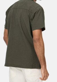Camisa de manga corta verde oliva vista desde atrás. Presenta un cuello estándar, un dobladillo limpio y una textura de algodón suave.